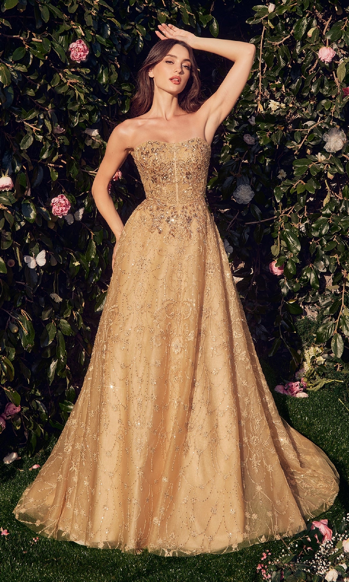 A woman stands outdoors amid greenery and pink flowers, wearing the Andrea & Leo MA104—a long sleeve, floor-length formal dress with intricate beading and embroidery.